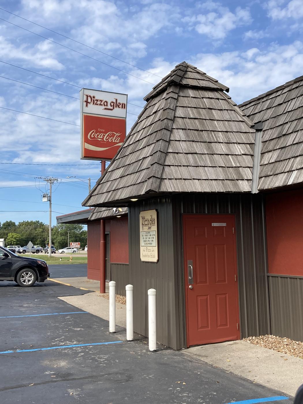 The front door of Pizza Glen in Clinton, Missouri, under a cedar shingle roof, with the hours board beside it